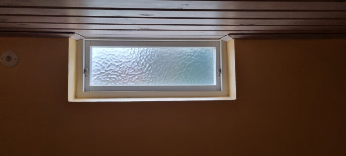 Liggande fönster med frostat glas mellan vita ramar, inbyggt i mörk vägg och ljus takbeklädnad.