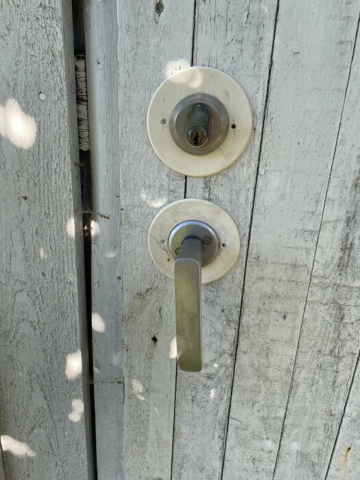 En vit sliten dörr med silverfärgat dörrhandtag och låscylinder, trästruktur synlig, solljus reflektioner.