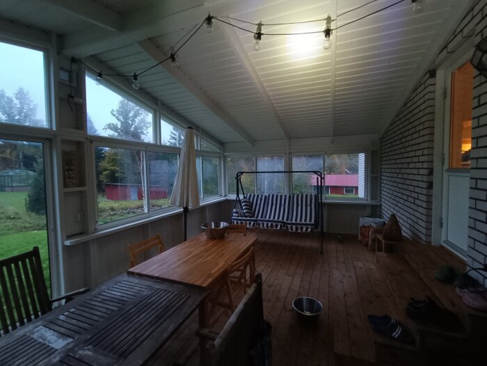 Inglasad veranda med trämöbler, soffa, fönster mot trädgård, kvällsljus, bekväm och hemtrevlig atmosfär.