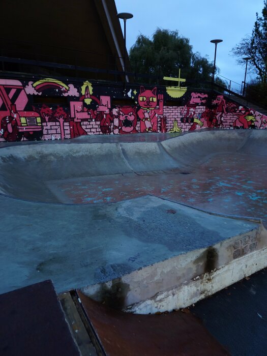 Tom skateboardpark med färgglada väggmålningar och betongramp mot kvällshimmel.