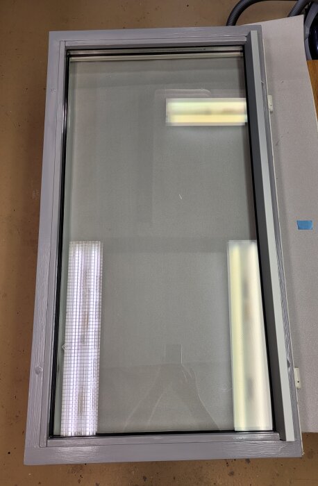 Vertikalt placerat fönster med reflektion av lysrör på en arbetsyta, grå ram och suddig bakgrund.