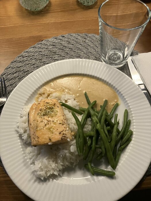 Middagsdukning med lax, ris, gröna bönor och vit sås. Tomt glas, grå duk, träbord.