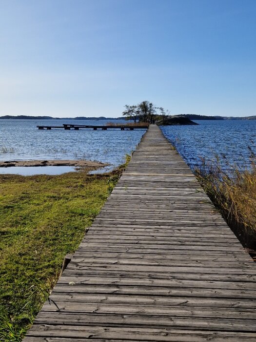 Träbrygga leder mot ö i sjö under klarblå himmel, bryggans slut ej synligt.