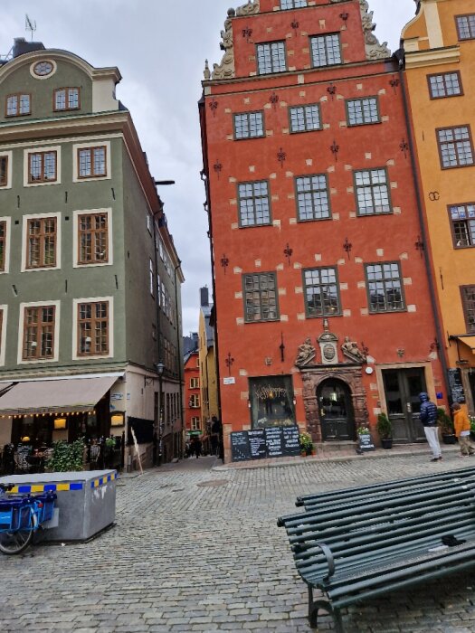 Gammelstadens kullerstensgata, historiska färgglada byggnader, uteservering, bänk, molnig himmel.