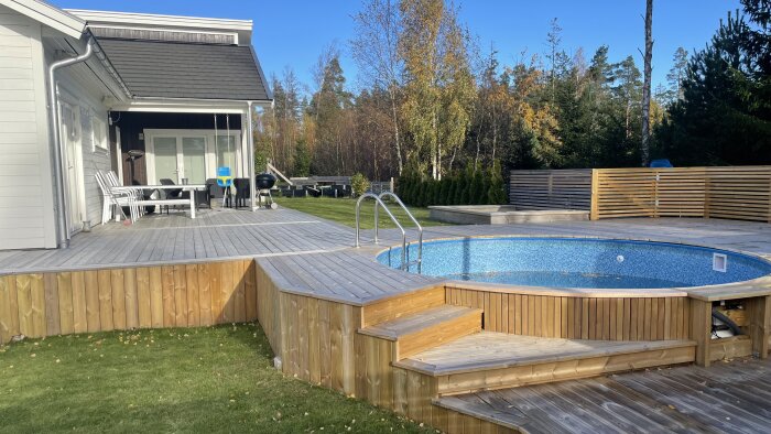 Hus med terrass, pool, trädäck, solstolar och grill, omgivet av träd och gräsmatta på dagen.