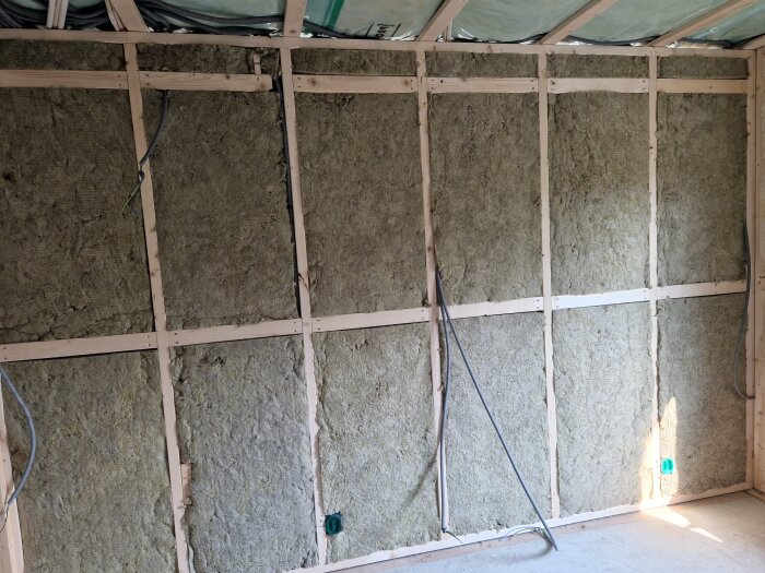 Inomhusvy av en vägg under konstruktion med träreglar och isoleringsmaterial, samt synliga elledningar.