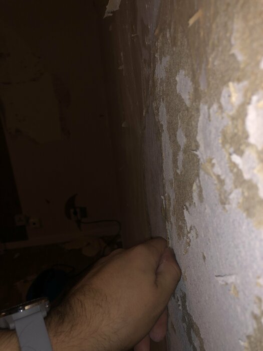 En hand skrapar tapet från en murad vägg; omgivning ser ut att vara i renovering.