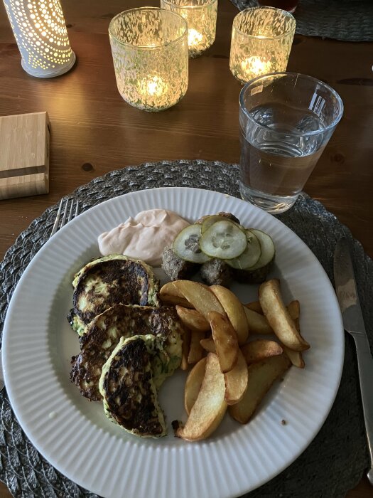 Middagsdukning med ljus, vatten, zucchiniplättar, klyftpotatis, sås och gurka på tallrik.