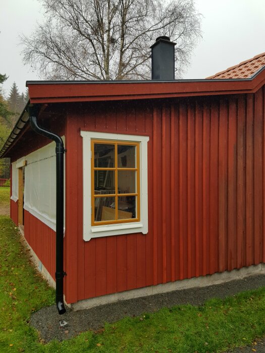 Rödfärgad träbyggnad, fönster med vita foder, svart stuprör, grå himmel, grönt gräs, regnigt väder.