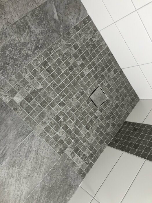 Hörn av ett modernt badrum med gråa, vita plattor och inbyggd golvbrunn.