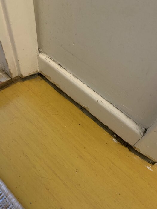 Hörn av ett rum med vit dörrkarm och gulaktigt golv, synliga slitage- och smutstecken.