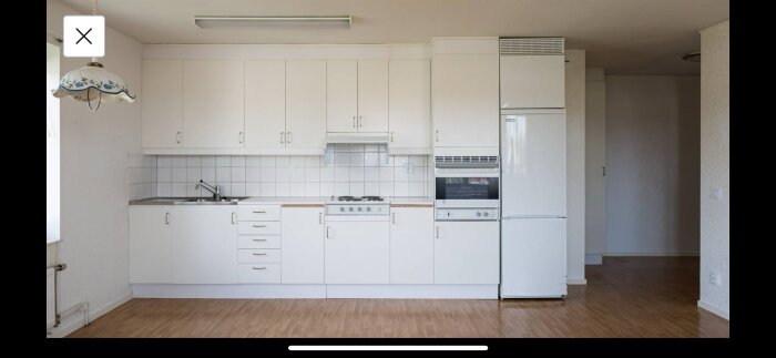 Modernt kök, vita skåp, spis, ugn, kylskåp, kakel, ljust golv, tom vägg, taklampa, neutrala färger.