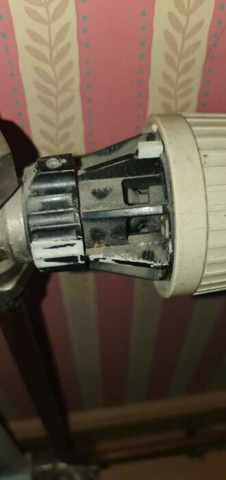 Delvis skadad, smutsig vit radiatortermostat mot mönstrad tapet, synlig mekanisk del, bär tecken på slitage eller skada.