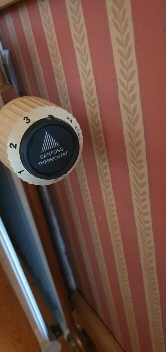 Danfoss termostat på en radiator, randig tapet i bakgrunden, inomhus, vinkel visar del av fönsterkarm.