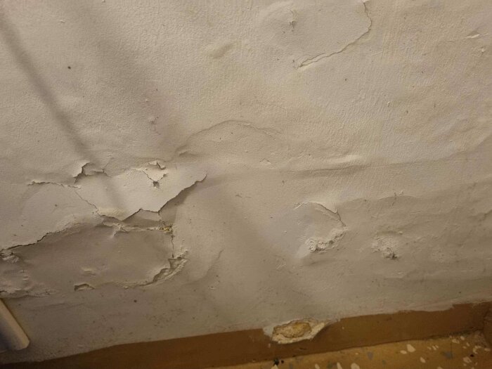 Vägg med flagnande färg och skador nära golvet, behöver reparation eller omålning.