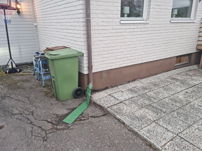 Husfasad, sopkärl, grönt spännband, stenplattor, fönster, korgställning, stege, sprickor i asfalten.
