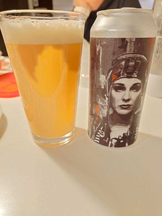 Ölglas med skum på bord bredvid konstnärlig ölburk, inomhus, delvis synlig person, ljus bakgrund.