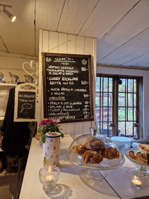 Kaféinteriör med tavla av lunchmeny, bakverk under glaskupa, ljus och blomma i vas.