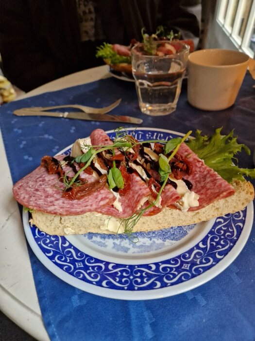 Smörgås med salami, ost, syltad lök på blåvit tallrik, vatten, kaffe, en suddig person i bakgrunden.