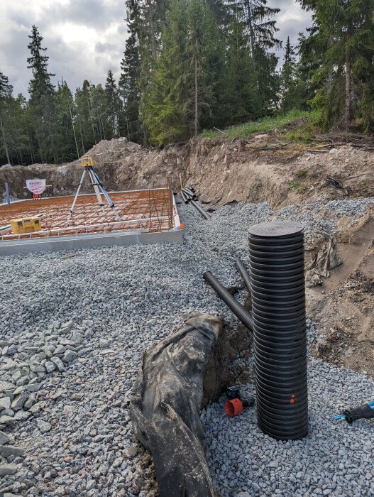 Byggplats med grus, armeringsjärn, rör, skog i bakgrunden, markarbete pågår.