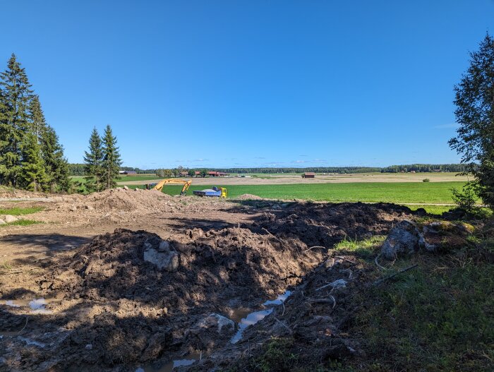 Markarbete, grävmaskin, gröna fält, skog, byggarbetsplats, soligt väder, blå himmel, ingen person synlig.