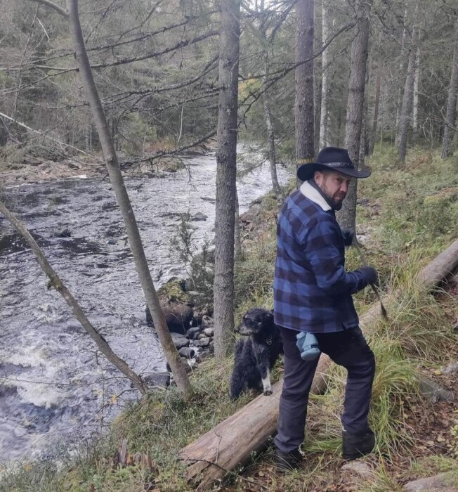 Man i hatt och skogskläder vid flod med hund, omgiven av träd och natur.