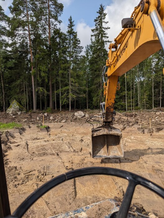 Grävmaskin på lerig mark, skog i bakgrunden, sikten från förarhytten, grävarm och skopa synlig.