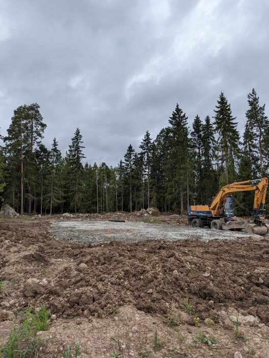 Byggplats i skog, grävskopa, röjning, grå himmel, jordhögar, ytberedning, skogsbakgrund.