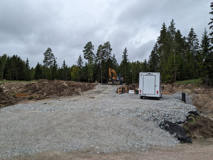 Byggarbetsplats i skogsmiljö, grävmaskin, släpvagn, grus, jordhögar, molnig himmel.