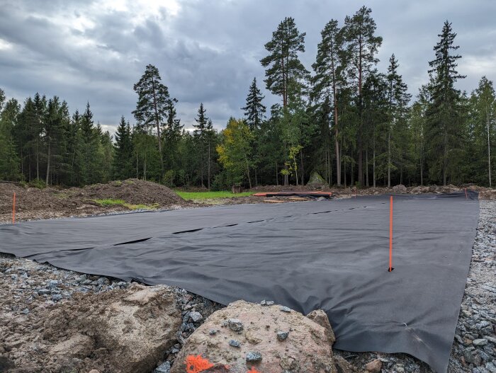 Tomt under förberedelse, mörk markduk, grushög, markeringsspikar, skogsbakgrund, mulet väder.