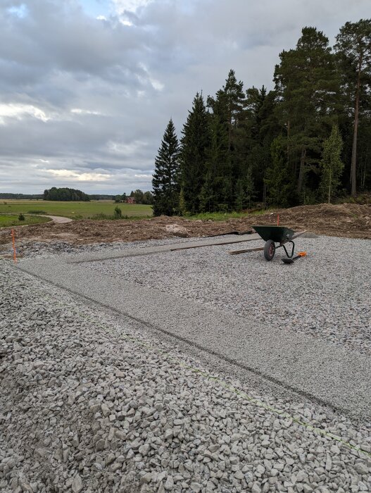 Grusgrund med skottkärra, skog bakom, öppet landskap i bakgrunden, molnig himmel.