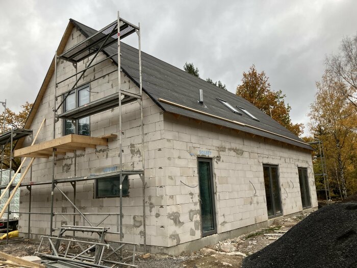 Tvåvåningshus under konstruktion med skal av betongblock, ställning, skiffer tak, höstträd i bakgrunden.