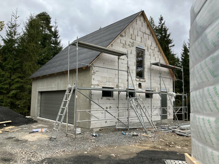 Ett hus under konstruktion med ställningar, stege och byggmaterial synliga. Skogsbakgrund och molnig himmel.