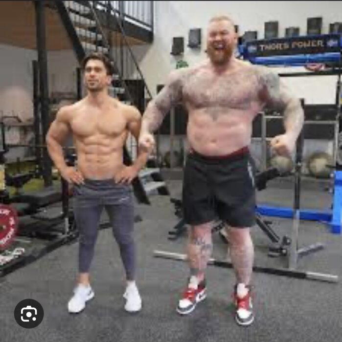 Två män visar muskler, en mycket större än den andre, i ett gym.
