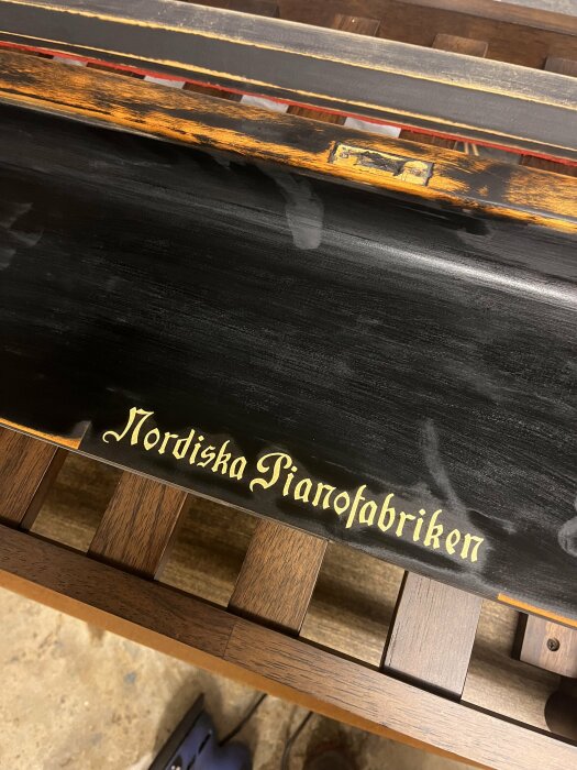 Ett svart piano med guldtext "Nordiska Pianofabriken" på fronten, synliga träslag, och nötta tangenter.