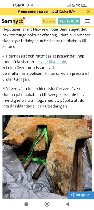 Ett stort, rostigt ankare med lyftstroppar på grå presenning.