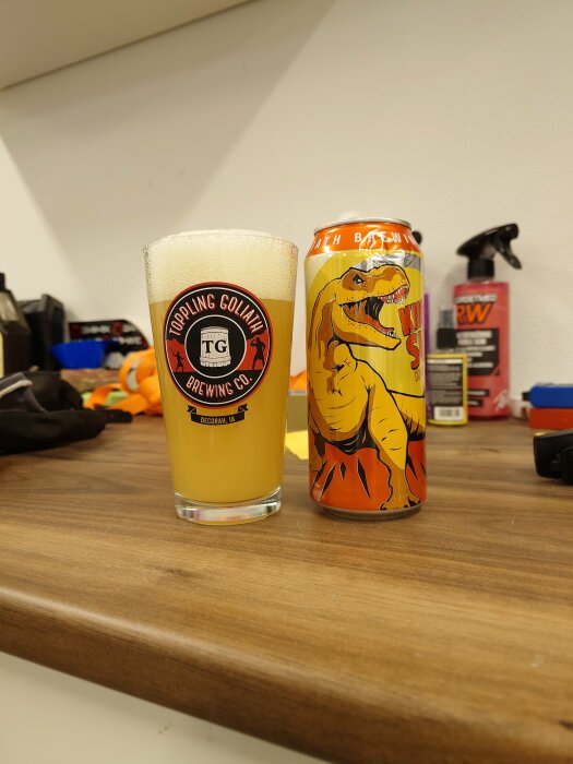 Ölglas med skum krona bredvid en dryckesburk med dinosaurie på arbetsbänk.