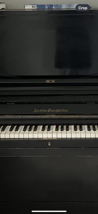 Svart upprätt piano. Tangenter synliga. Logotyp: "Nordiska Pianofabriken". Beskärningsoption överst. Inredningselement.