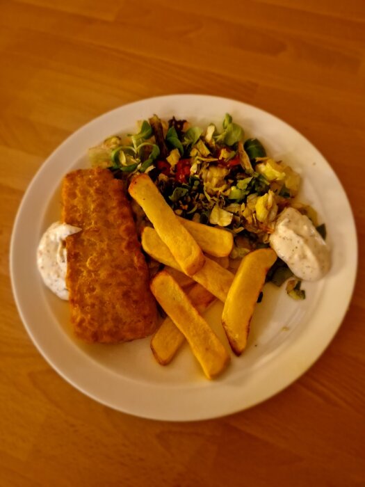 Ett måltidsfat med fisk, pommes frites, grönsallad och vit sås på träbord.