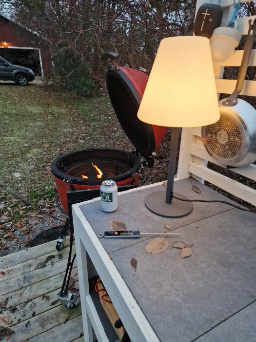 Utomhuskök med öppen grill, ölburk, fällkniv, höstlöv, och bordslampa i skymningen.