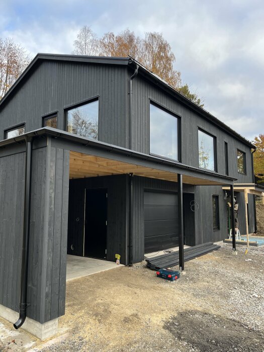 Modernt svart hus med stora fönster och carport under konstruktion, omgiven av grus och natur.