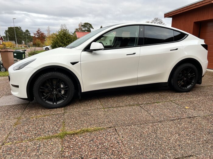 Vit elbil parkerad utanför hus, sannolikt Tesla Model Y, med svarta fälgar på uppfart.