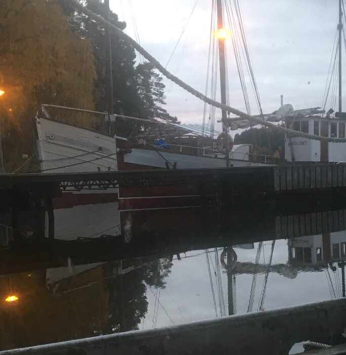 Träsegelbåtar vid kaj, spegling i vatten, kvällsljus, lugnt, marinmiljö, mast, höstträd, gatubelysning.