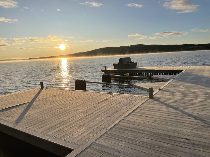 Solnedgång vid sjö, brygga, bänk, dimma, solens reflexion på vatten, fridfullt.