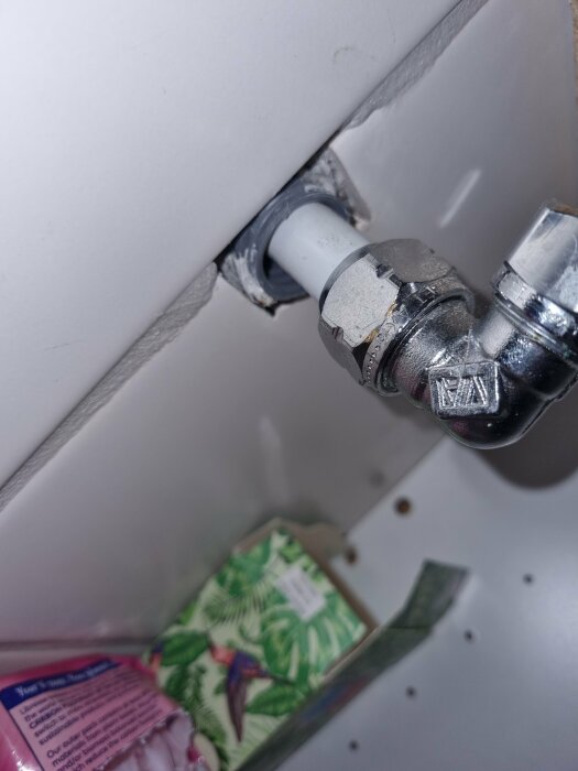 Vattenrör genom vägg, silverfärgad ventil, ojämn hålighetsförsegling, vit bakgrund, delvis synligt skåp.