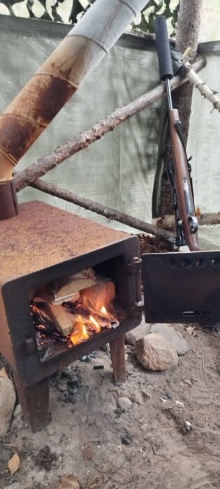 Rostig kamin i utomhusmiljö med öppen eld, stödd av stenar och en gevär står lutat intill.
