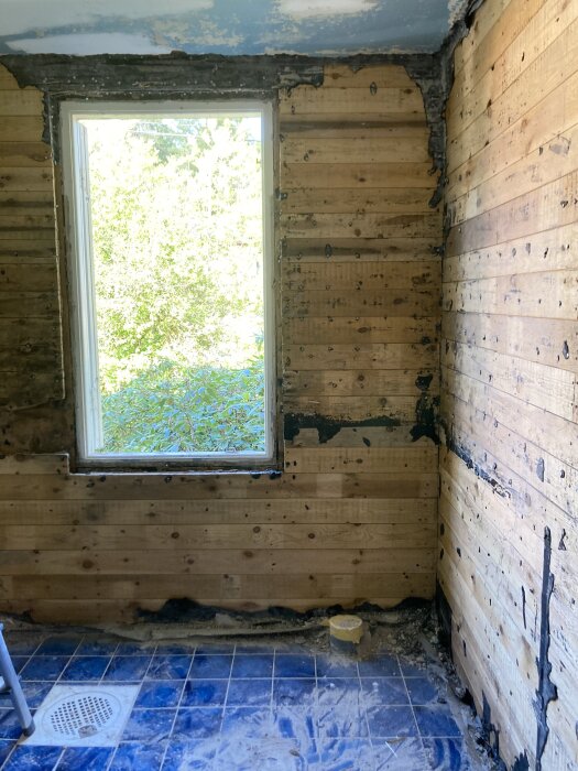 Ett renoveringsprojekt. Rum med nakna trästommarväggar, ett fönster, blått golv, avlägsnat innertak.