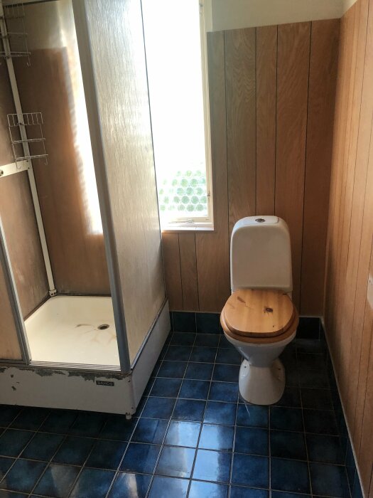 Ett badrum med duschhörna, toalett och träpaneler. Blå kakel på golvet. Naturligt ljus från fönstret.
