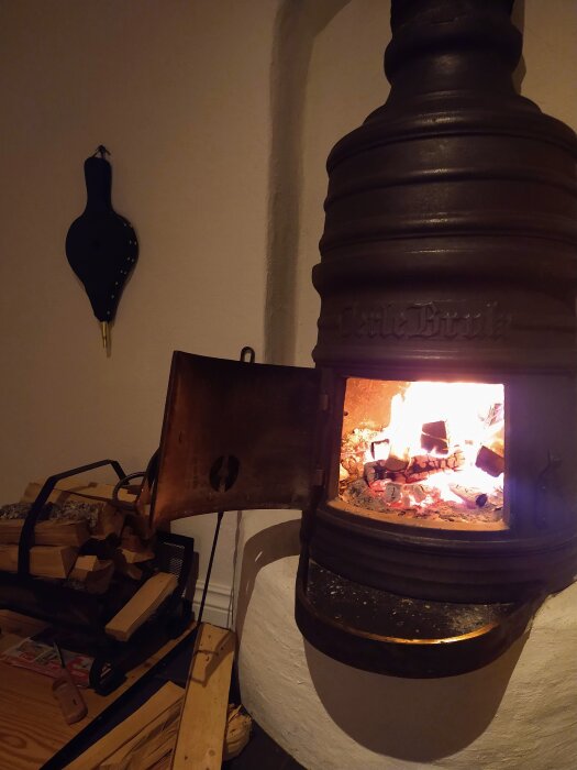 En äldre gjutjärnskamin brinner med öppen lucka, omgiven av ved, i ett mysigt hemmiljö. Väggklocka syns också.