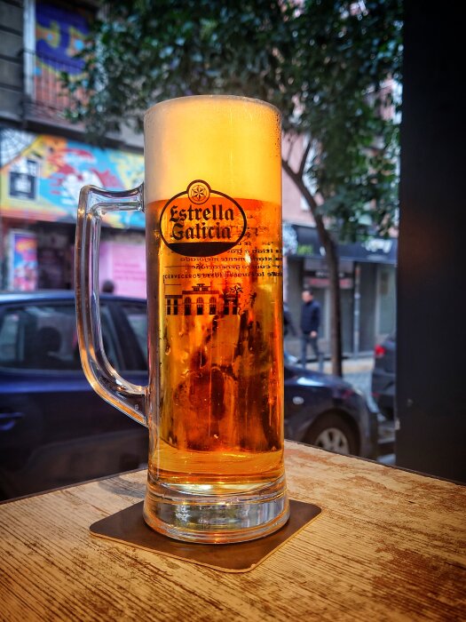 Ett stort glas öl på träbord, Estrella Galicia-logotype, citymiljö i bakgrunden.